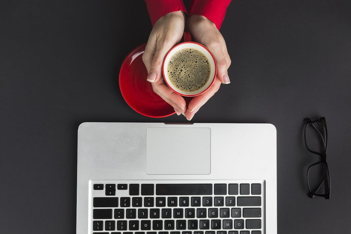 Imagem de um notebook aberto e duas mãos segurando um café a sua frente