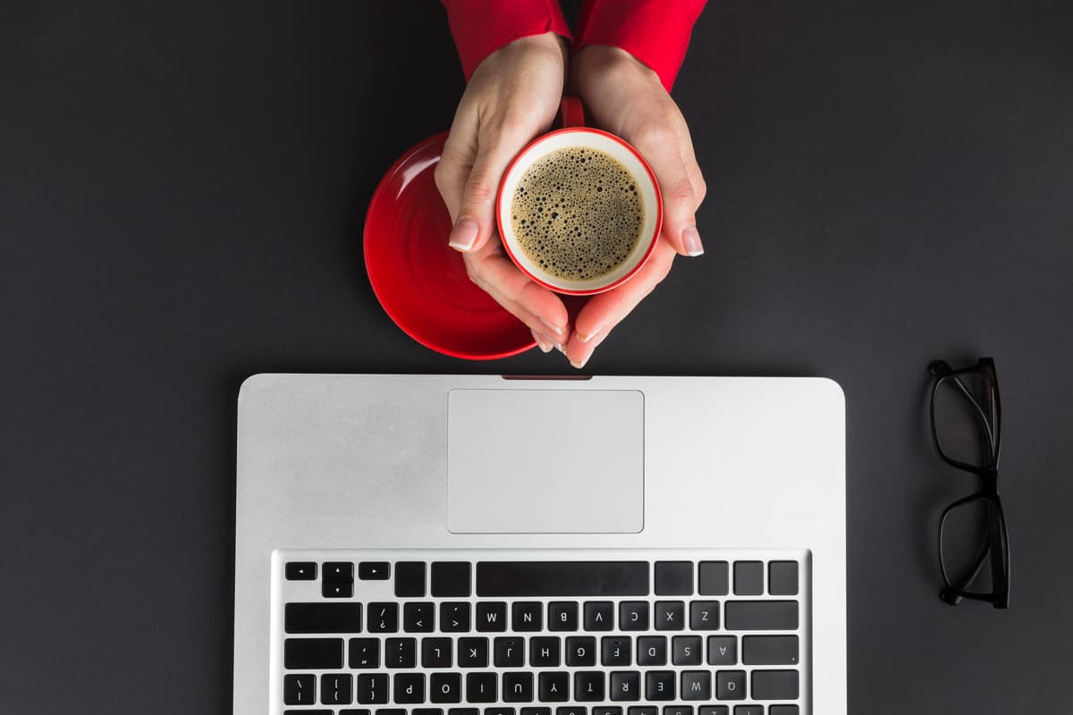 Imagem de um notebook aberto e duas mãos segurando um café a sua frente